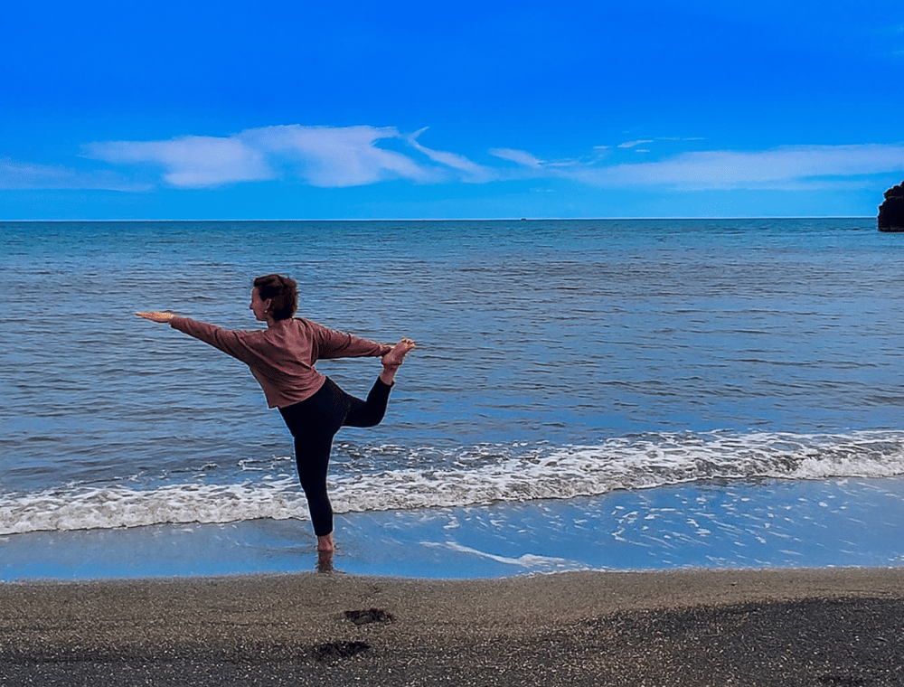 Héloïse Peyronnet pratiquant une posture de yoga sur une plage, debout au bord de l’eau, une jambe levée vers l’arrière et les bras étendus. La mer est calme et le ciel légèrement nuageux, avec deux rochers visibles à l’horizon.